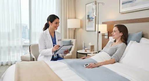 Doctor with a tablet talking with a woman in a hotel bed wearing a pulse oximeter, IV stand nearby