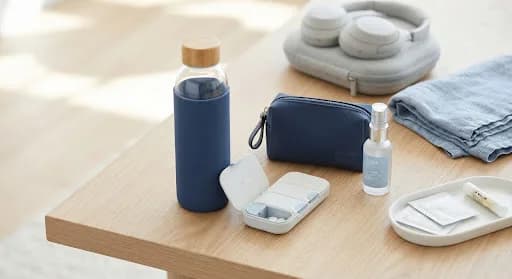 Travel essentials on a wooden table: water bottle, pill organiser, zip pouch, spray bottle and headphones